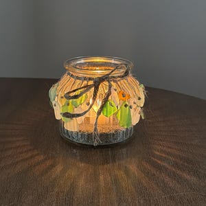 May include: A glass candle holder with a warm glow. The jar is adorned with green, white, and orange decorative elements and tied with twine. The candle holder is sitting on a dark brown surface.
