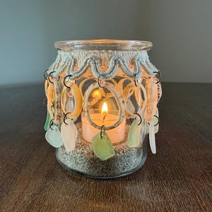 May include: A clear glass candle holder filled with sand and a lit tea light. The rim is wrapped in cream-colored macrame cord, from which hang various small seashells and smooth pieces of sea glass in shades of green, white, and tan.
