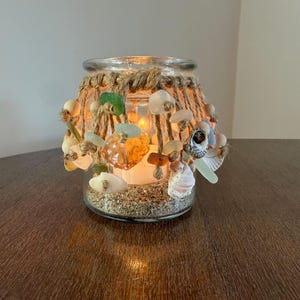 Pode incluir: Um suporte de vela de vidro transparente preenchido com areia na base e envolto em uma rede de corda de juta natural. A rede é decorada com várias conchas pequenas, pedaços de vidro do mar polido em verde e âmbar e contas de madeira clara.