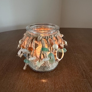 May include: A clear glass candle holder filled with sand and a lit tea light. The rim is wrapped in thick jute twine with hanging strands decorated with smooth sea glass pieces in shades of green and white, wooden beads, and a small seashell.