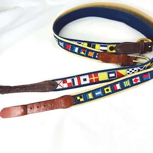 May include: Two nautical-themed belts with brown leather accents and brass buckles. The belts feature a navy blue and tan canvas design with colorful signal flags. One belt is partially buckled, showcasing the leather and buckle details.