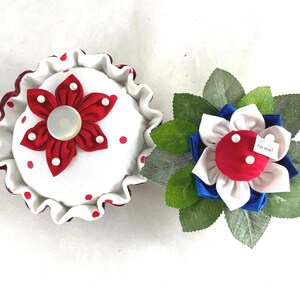 May include: Two pincushions, one shaped like a cupcake with red and white polka dots and a red flower, the other shaped like a flower with red, white, and blue petals and green leaves. The flower pincushion has a tag that says "Pin me!"