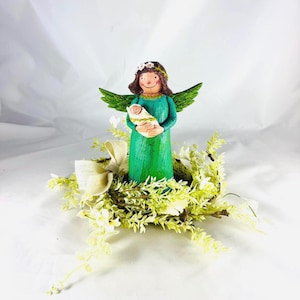 May include: A handcrafted angel figurine in a green dress, holding a baby wrapped in a white blanket. The angel has green wings and a floral crown. The figurine is surrounded by a wreath of white and green foliage.
