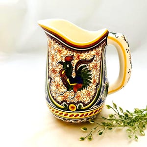 May include: A ceramic pitcher with a rooster design. The pitcher features a cream-colored interior and a handle with yellow and black accents. The exterior is decorated with a rooster, floral patterns, and a border of yellow, brown, and blue.