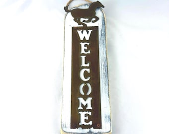 May include: Wooden welcome sign with a distressed white and brown finish. The sign features a cutout metal panel with the word "WELCOME" and a horse silhouette at the top. A jute rope is attached for hanging.
