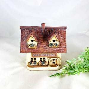 May include: A ceramic house figurine with a textured brown shingled roof and cream-colored walls. The front features two dormer windows with heart-shaped cutouts, flower boxes, and a sign above the ground floor windows that reads KEEP TRYST INN.