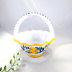 May include: White ceramic basket with a handle, featuring a scalloped rim and a yellow base. The basket is adorned with blue and yellow floral patterns. The handle has blue accents.