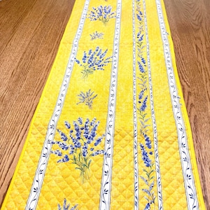 May include: Yellow quilted table runner with a repeating pattern of blue lavender bouquets and vertical white stripes with a leaf design. The runner is placed on a wooden table and adds a touch of color and pattern to the space.