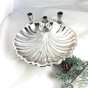 May include: A polished silver-toned metal dish shaped like a scallop shell, featuring three vertical cylindrical candle holders attached to the top edge. The shell design includes radiating fluted grooves and a decorative scroll detail at the center.