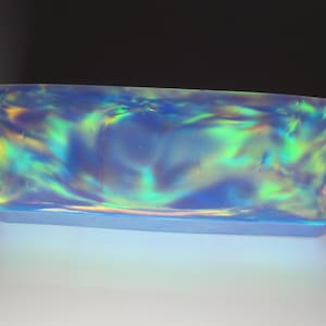 Peut inclure: Objet rectangulaire en verre irisé avec une base bleu clair et un motif tourbillonnant de bleus, de verts et de jaunes. L'objet a des coins arrondis et une surface lisse et réfléchissante. L'effet global est un jeu de lumière et de couleur.