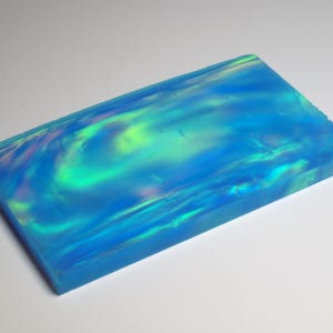 Peut inclure: Une plaque rectangulaire en opale synthétique présentant une couleur de base bleu vif avec des motifs tourbillonnants de reflets verts et rose clair irisés qui se déplacent sur la surface polie et semi-translucide du bloc plat.