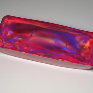 Peut inclure: Un bloc rectangulaire à la surface lisse et polie, présentant une palette de couleurs irisées vibrantes de rose profond, magenta, avec des éclats de bleu électrique et de violet qui se déplacent sur le matériau translucide en vagues.