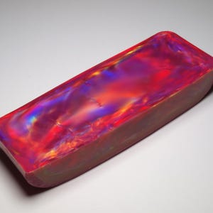 Peut inclure: Un bloc rectangulaire à la surface lisse et polie, présentant un motif tourbillonnant vibrant de teintes rouge profond, magenta et violet irisé qui changent d'intensité sur la face supérieure, posé sur une base solide de couleur claire.
