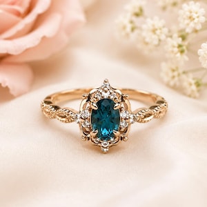 Könnte beinhalten: Ein goldener Ring mit einem oval geschliffenen, dunkelblauen Edelstein in der Mitte, umgeben von einem Kranz aus kleinen, klaren Steinen. Die Ringschiene hat ein filigranes, strukturiertes Design mit weiteren kleinen, klaren Steinen.