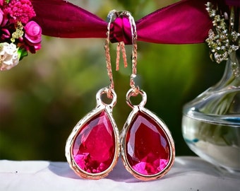 May include: A pair of teardrop-shaped earrings with a vibrant red gemstone set in a gold-colored metal. The earrings hang from delicate gold chains. The background features a blurred green and white floral arrangement.