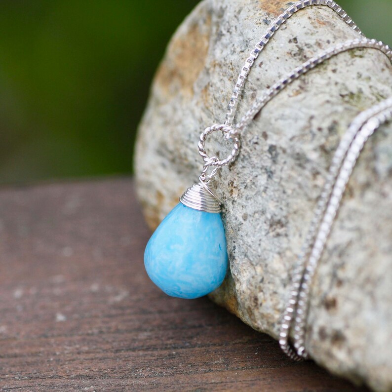 May include: A silver chain necklace with a light blue teardrop-shaped pendant. The pendant is wrapped in silver wire.