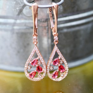 May include: Rose gold dangle earrings with a teardrop design. The earrings are adorned with clear and red cubic zirconia stones.