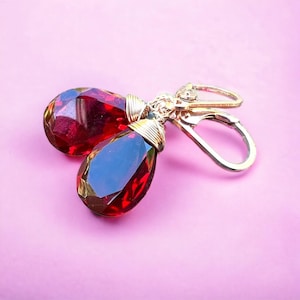 May include: A pair of teardrop earrings featuring faceted, deep red gemstones. The gems are set in gold-coloured metal with a wire-wrapped design at the top, and gold-coloured lever-back closures. The background is a solid, light pink colour.