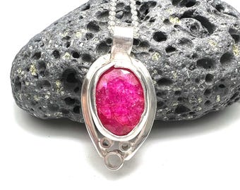May include: A silver pendant necklace with a large, oval-shaped, hot pink gemstone. The gemstone is set in a silver bezel with a delicate chain. The pendant is displayed against a dark, textured background.