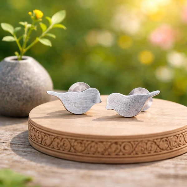 Pode incluir: Dois brincos prateados em forma de pássaro são exibidos em um suporte circular de madeira. Os brincos têm uma textura de metal escovado e estão sobre um fundo verde e amarelo desfocado. Um pequeno vaso com uma planta está ao fundo.