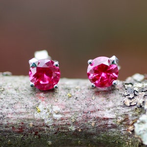 May include: A pair of stud earrings featuring round, faceted, hot pink gemstones set in silver-colored metal. The earrings are displayed on a natural, textured wooden surface, with a blurred background of muted greens and browns.