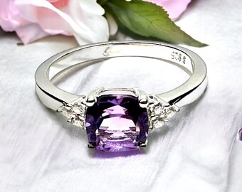 May include: A silver ring with a large, square-shaped amethyst gemstone surrounded by smaller, clear gemstones. The ring is set on a white background.
