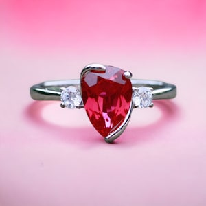 May include: A silver ring featuring a large, pear-shaped red gemstone and two smaller, round clear gemstones.