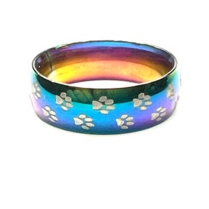 Peut inclure: Une bague en acier inoxydable aux couleurs de l'arc-en-ciel avec un motif de patte. La bague est une bande large avec une finition lisse et polie.