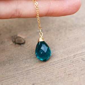 K&ouml;nnte beinhalten: Eine goldfarbene Halskette mit einem facettierten, tropfenf&ouml;rmigen Anh&auml;nger in Teal. Der Anh&auml;nger ist etwa 2 cm lang und oben mit Draht umwickelt. Die Halskette wird vor einem Holzuntergrund pr&auml;sentiert.