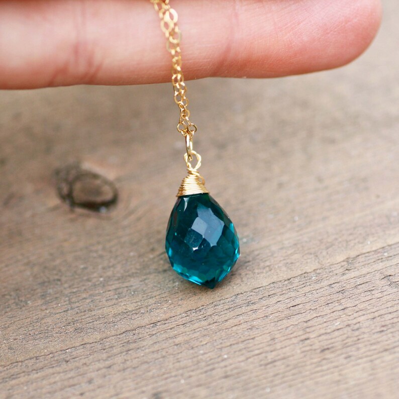 K&ouml;nnte beinhalten: Eine goldfarbene Halskette mit einem facettierten, tropfenf&ouml;rmigen Anh&auml;nger in Teal. Der Anh&auml;nger ist etwa 2 cm lang und oben mit Draht umwickelt. Die Halskette wird vor einem Holzuntergrund pr&auml;sentiert.
