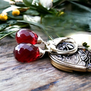 May include: A pair of earrings with red gemstone drops and gold wire accents. The earrings have silver leverback closures.
