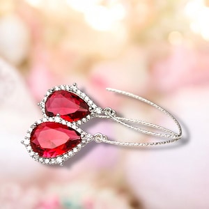 May include: A pair of elegant drop earrings featuring large, teardrop-shaped red gemstones. Each gem is surrounded by a halo of small, sparkling white stones, set in a silver-toned metal. The earrings have a delicate, curved design.