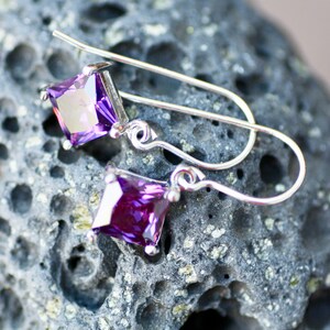 May include: A pair of silver earrings with purple square-shaped gemstones. The earrings are on a textured gray surface.
