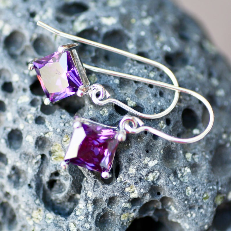May include: A pair of silver earrings with purple square-shaped gemstones. The earrings are on a textured gray surface.