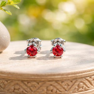 May include: A pair of stud earrings with bright red gemstones. The earrings have a silver setting with small accent stones around the main stone. The background is a soft focus of green foliage.