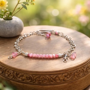 May include: A delicate silver bracelet featuring a strand of pink gemstones, accented with smaller pink charms and white beads. The bracelet is displayed on a wooden surface, with a blurred background of greenery and flowers. The bracelet is approximately 7 inches in length.