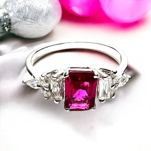 May include: A silver ring with a large, emerald-cut ruby gemstone surrounded by smaller, pear-shaped diamonds.