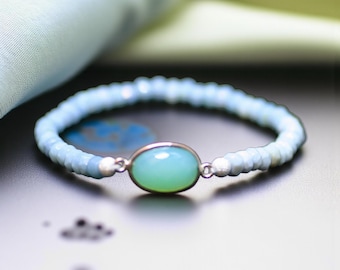 May include: A light blue beaded bracelet with a central oval-shaped, light green stone set in a silver-colored metal. The bracelet is on a dark surface, with a soft, light blue fabric in the background.