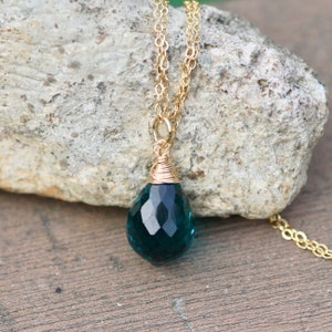 K&ouml;nnte beinhalten: Eine goldfarbene Halskette mit einem facettierten, tropfenf&ouml;rmigen Anh&auml;nger in Teal. Der Anh&auml;nger ist mit goldfarbenem Draht umwickelt. Die Halskette wird auf einer grauen Felsfl&auml;che pr&auml;sentiert.