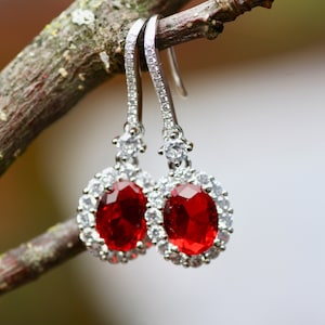 May include: A pair of silver dangle earrings with a red oval gemstone surrounded by a halo of smaller clear gemstones.