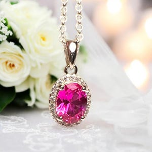 May include: A silver necklace featuring a pink oval gemstone pendant, encircled by small, clear stones. The backdrop includes white roses and soft lighting, creating a delicate aesthetic.