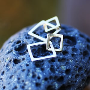May include: Silver geometric earrings with a square shape. The earrings are stacked on top of each other and are on a dark grey textured surface.