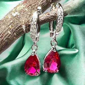 Puede incluir: Un par de pendientes de plata con piedras preciosas rojas en forma de lágrima. Los pendientes tienen un aro de plata con una fila de pequeñas piedras transparentes.