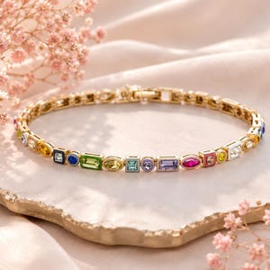 May include: A gold bracelet with a series of colourful, geometric gemstones. The gemstones are in various shapes, including squares, rectangles, and ovals, and are set in a delicate chain. The bracelet is displayed on a light-coloured surface with pink flowers.