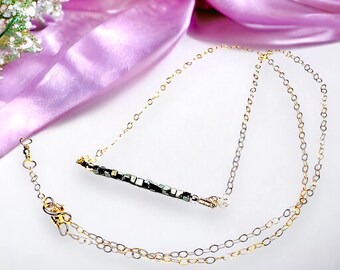 Op de afbeelding: Een delicate goudkleurige ketting met een staafhanger met kleine, donkergekleurde rechthoekige kralen. De ketting heeft een dubbele kettingontwerp en een karabijnsluiting. Een zacht, paars lint en witte bloemen staan op de achtergrond.