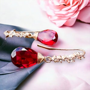 May include: A pair of gold earrings with red teardrop-shaped gemstones and clear crystal accents.