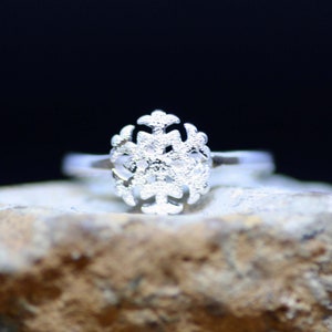 Puede incluir: Un anillo de plata con un diseño de copo de nieve. El anillo está sobre una roca.