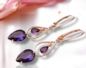 May include: A pair of rose gold earrings with a teardrop-shaped, deep purple gemstone. Each earring features a smaller, triangular purple stone and a lever-back closure. The earrings are displayed on a white surface.