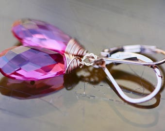 May include: A pair of drop earrings featuring faceted, teardrop-shaped pink gemstones. The gems are wire-wrapped in a copper-colored metal and attached to silver-colored lever-back closures. The earrings are displayed on a reflective surface.