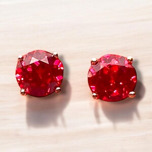 May include: Two round, faceted red gemstone stud earrings. The gems are set in gold-coloured metal with four prongs. The earrings are displayed on a light-coloured wooden surface.